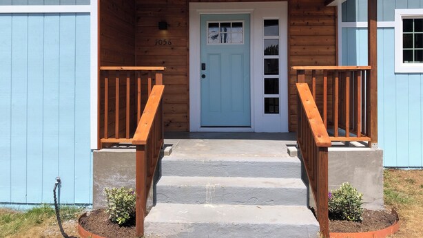 A wooden house with a light blue door, numbered 1056, and a concrete staircase leading to a porch with wooden railings.
