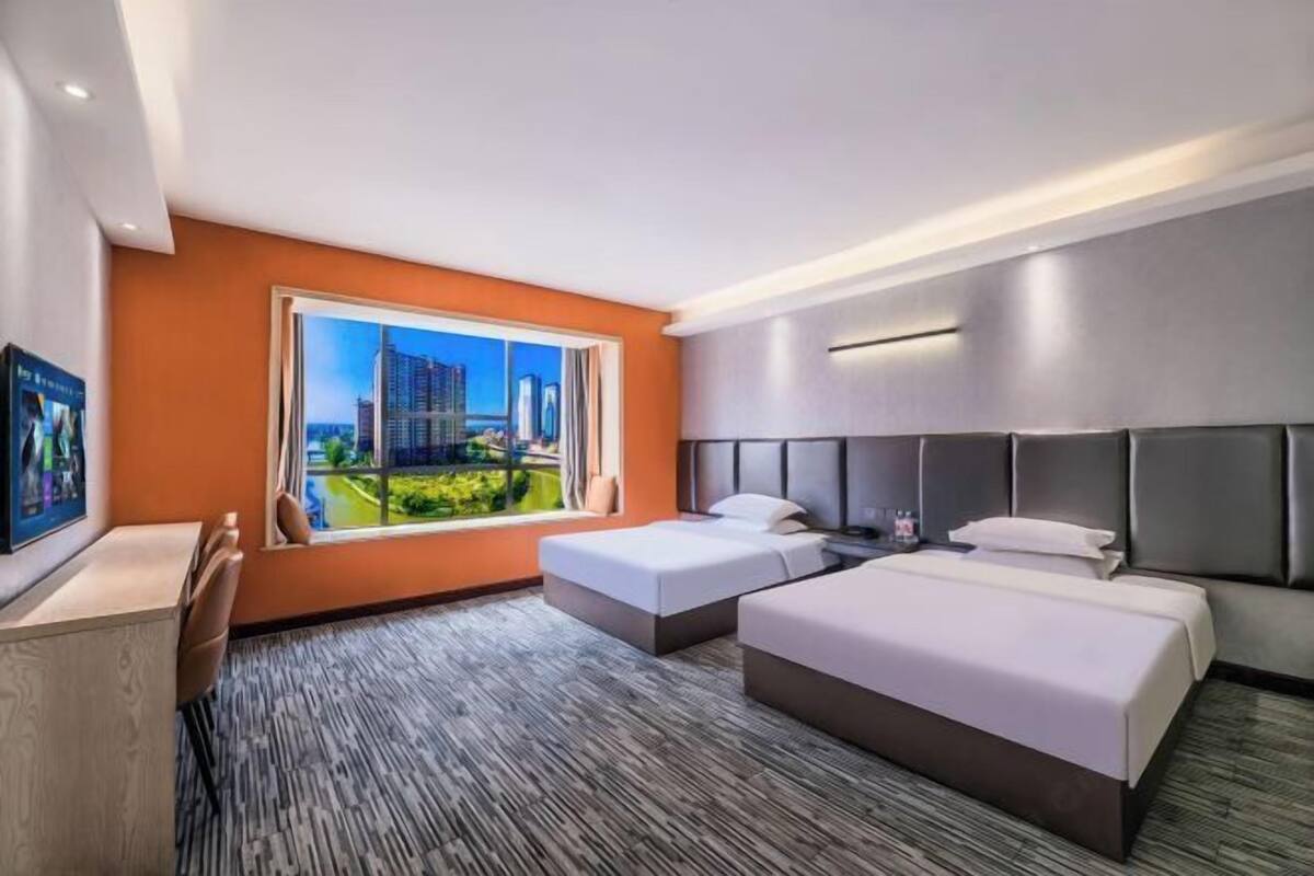 Ein Hotelzimmer mit zwei Betten, einem großen Fenster mit Blick auf die Stadt, einem Schreibtisch und einem Fernseher.