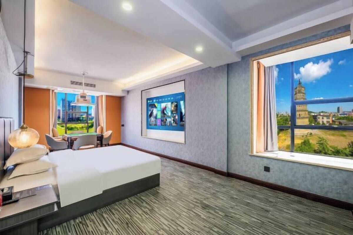 Ein Hotelzimmer mit Bett, Schreibtisch, Stuhl, Lampe, einem gerahmten Bild und einem großen Fenster mit Blick auf einen Turm.