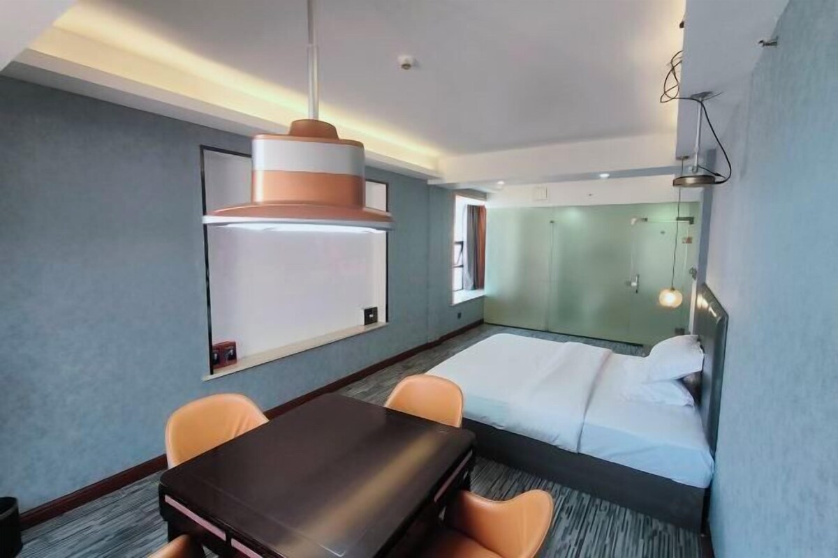 A hotel room with a bed, a desk, two chairs, a large pendant light, a whiteboard, and a glass partition.
