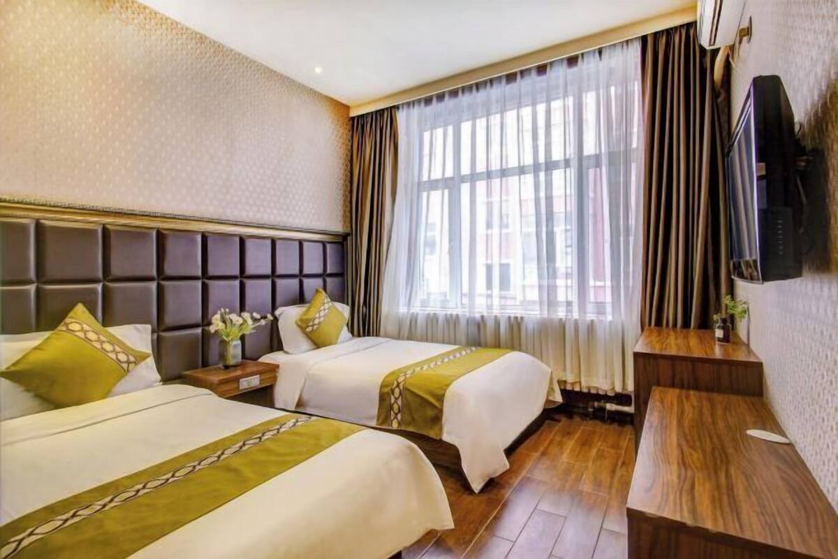 A hotel room with two beds, a wooden desk, a television, and a large window with curtains.