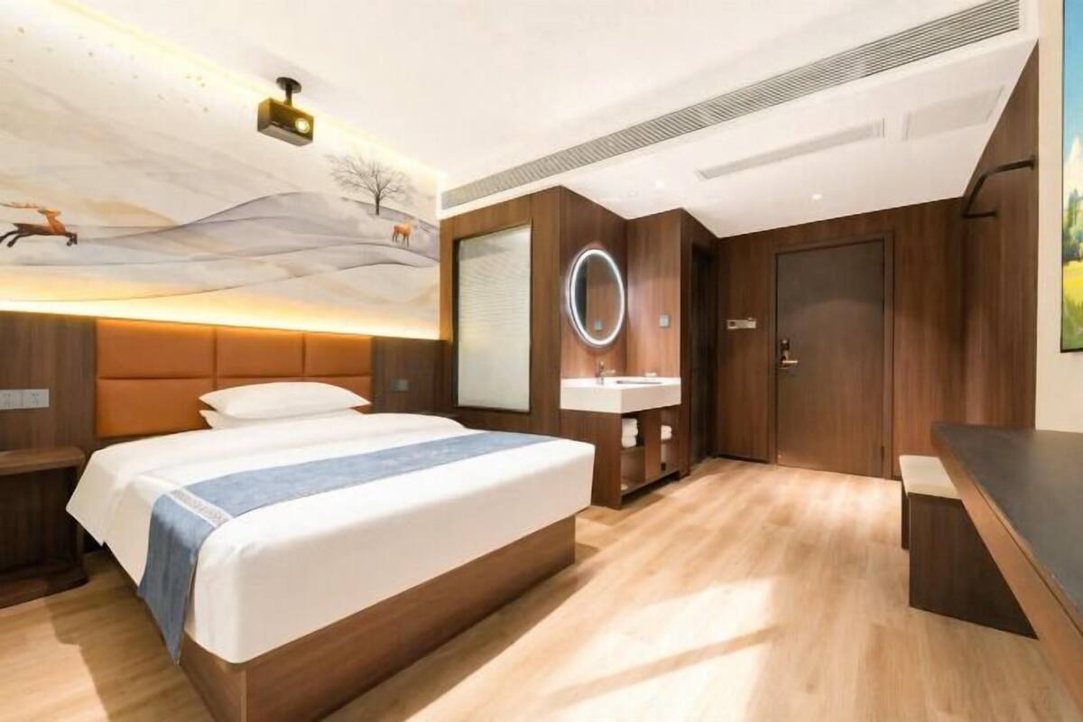 A modern hotel room with a large bed, a bedside table, a built-in desk with a mirror, and a bathroom with a sink and mirror.