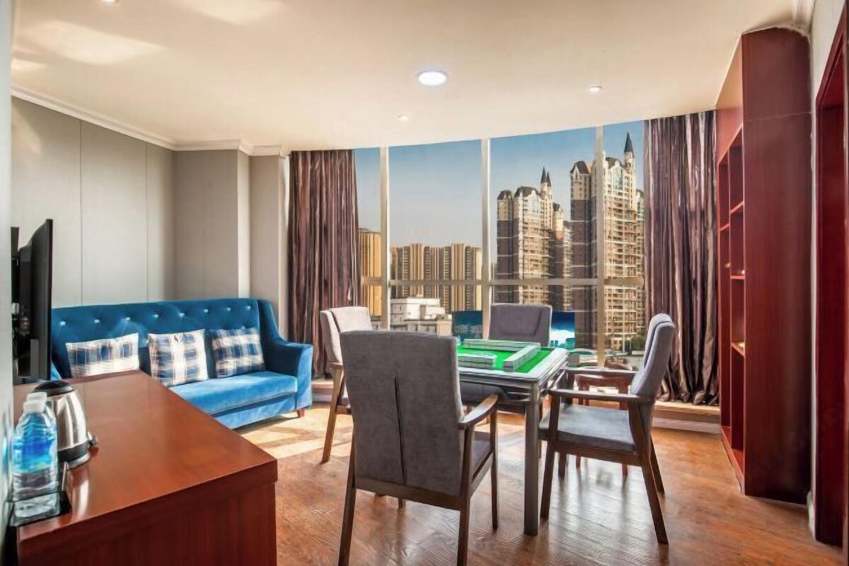 A modern hotel room with a city view, a blue sofa, a wooden desk, and a gaming table.