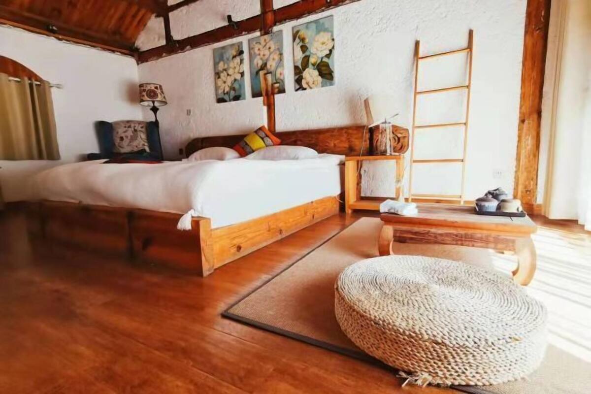 A wooden bed with a headboard, a bedside table with a lamp, a woven pouf, and a ladder against the wall.