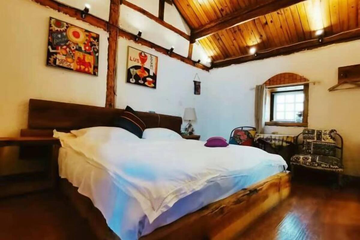A room with a wooden ceiling, a bed with white bedding, a chair, and artwork on the walls.