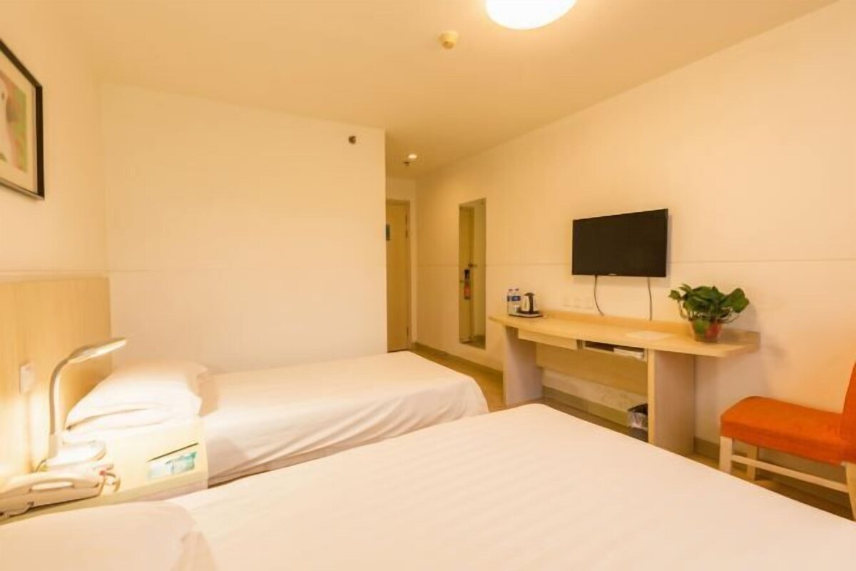 A hotel room with two beds, a desk with a flat-screen TV, a chair, and a bedside table with a lamp.