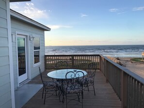 Eine Holzterrasse mit Tisch und Stühlen aus Glas, mit Blick auf Strand und Meer.
