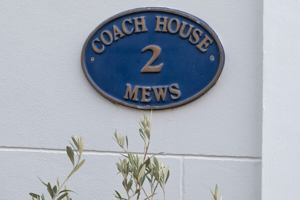 En blå husnummerplade med teksten "COACH HOUSE 2 MEWS" på en hvid væg.