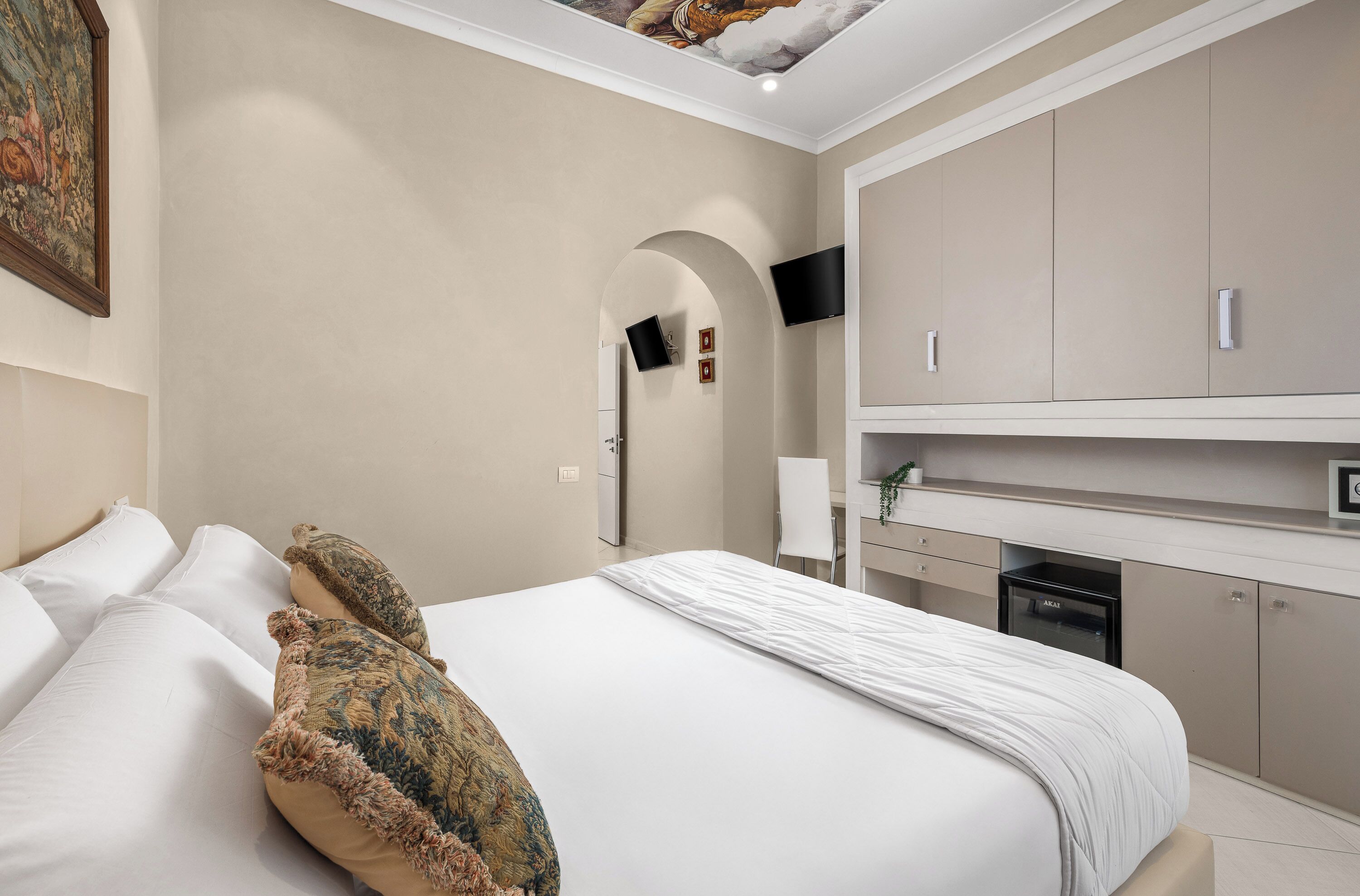 A bedroom with a bed, pillows, a TV mounted on the wall, a desk with a chair, and a built-in oven.