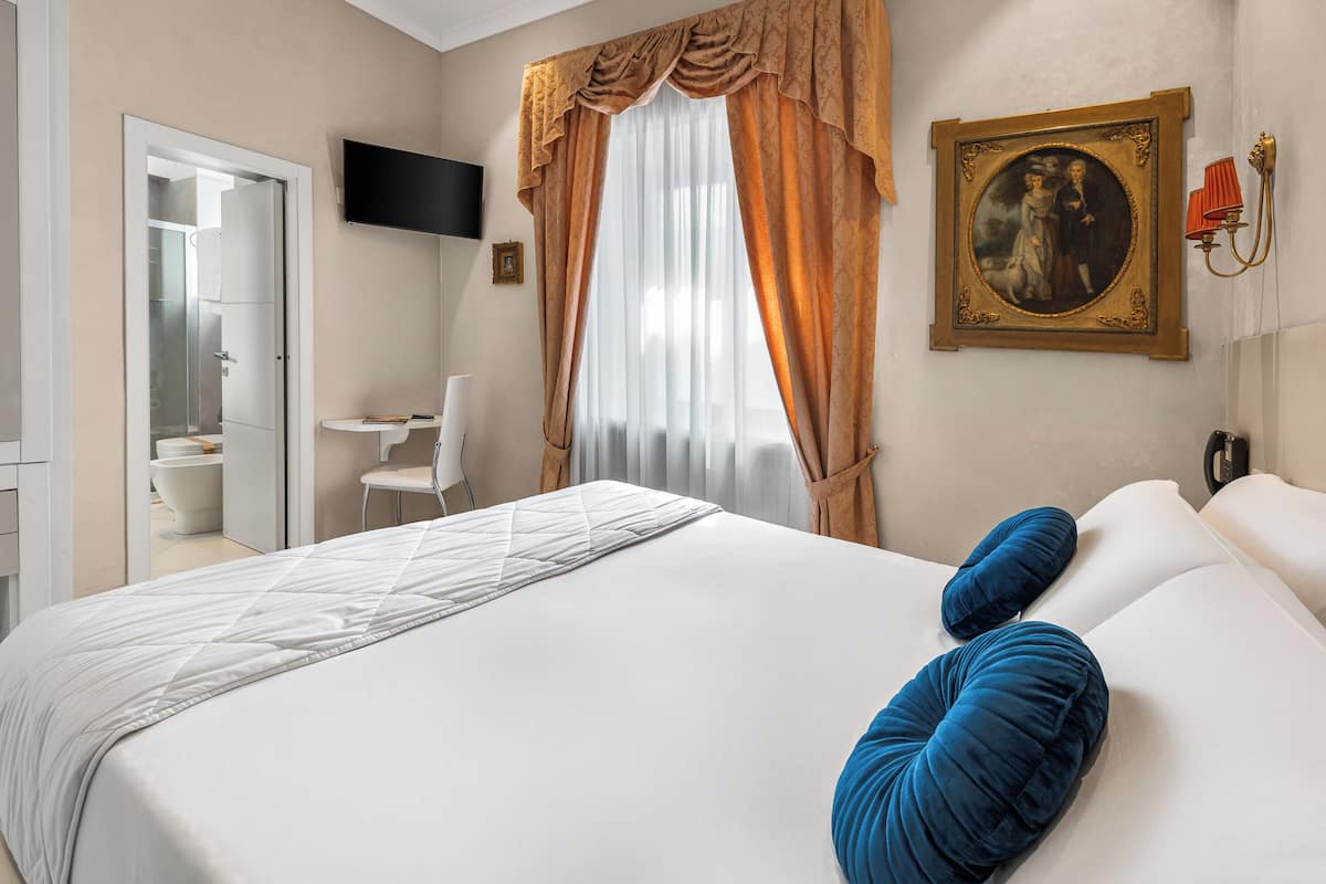 Une chambre d’hôtel avec un lit, une télévision, un tableau et une salle de bain visible à travers une porte ouverte.