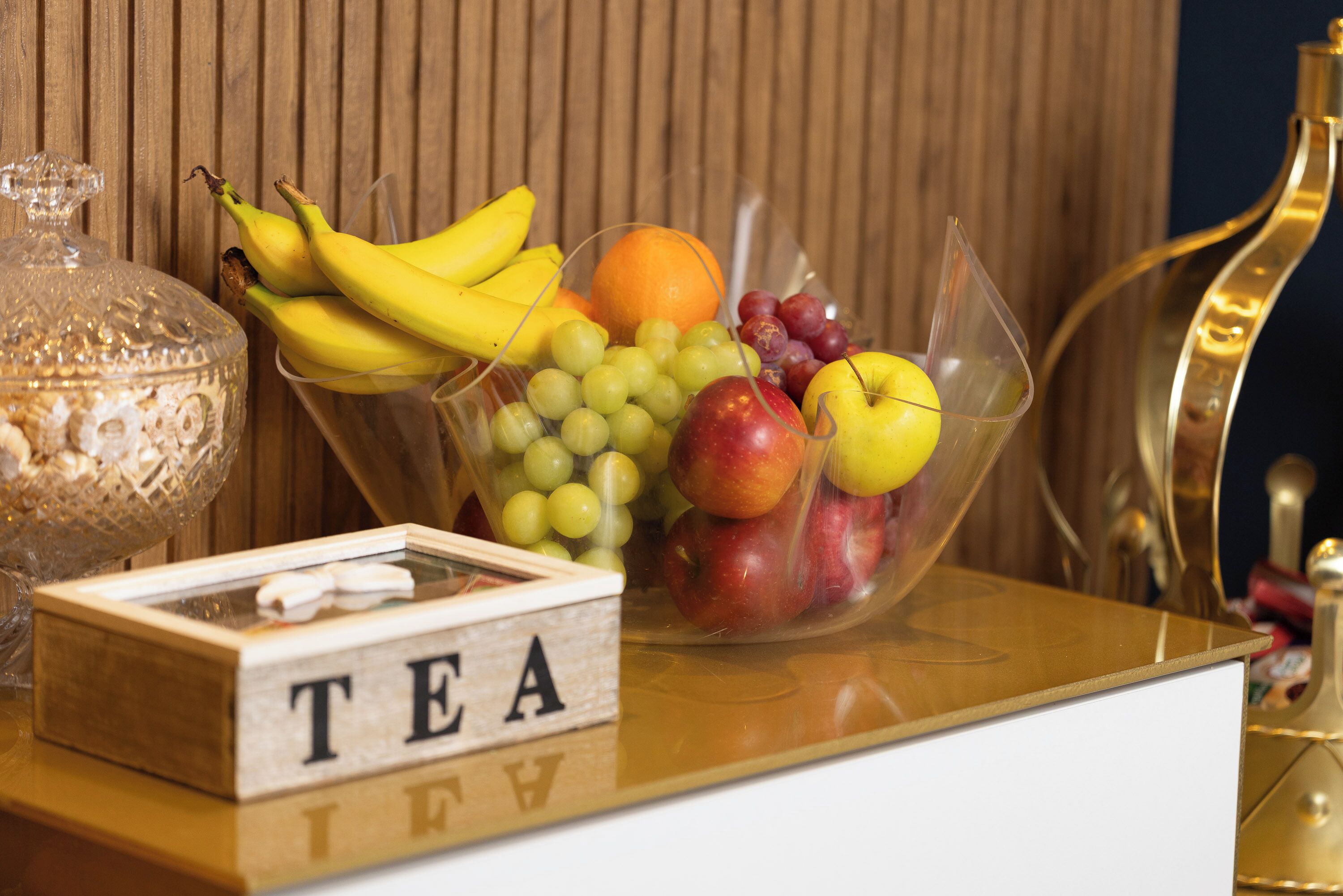 A fruit bowl with bananas, grapes, apples, and an orange on a table with a 'TEA' sign.