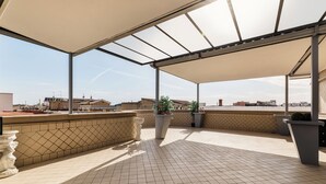 Een overdekt terras buiten met betegelde vloer, plantenpotten en uitzicht op omliggende gebouwen.