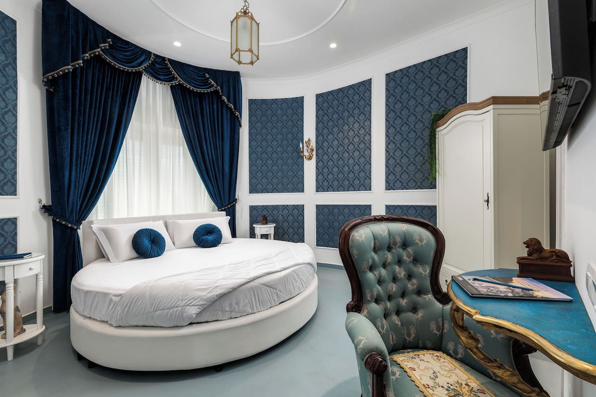 Une chambre à coucher avec un lit rond, un fauteuil bleu recouvert de tissu, une petite table et un grand miroir.