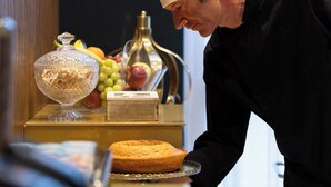 Een kok in een zwarte uniform en hoed, die een schaal met taart vasthoudt, in een keuken met een fruitschaal en een fles op het aanrecht.