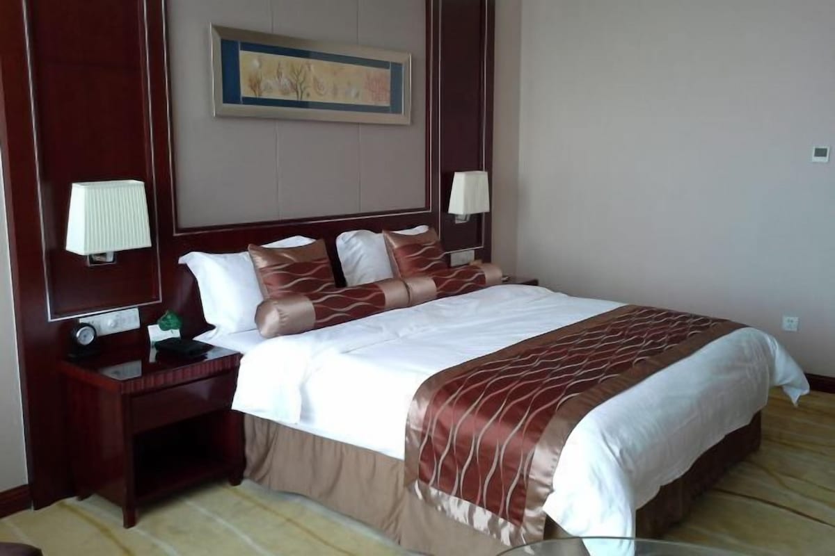 A hotel room with a large bed, two bedside tables, a nightstand, a wall-mounted lamp, and a framed picture on the wall.