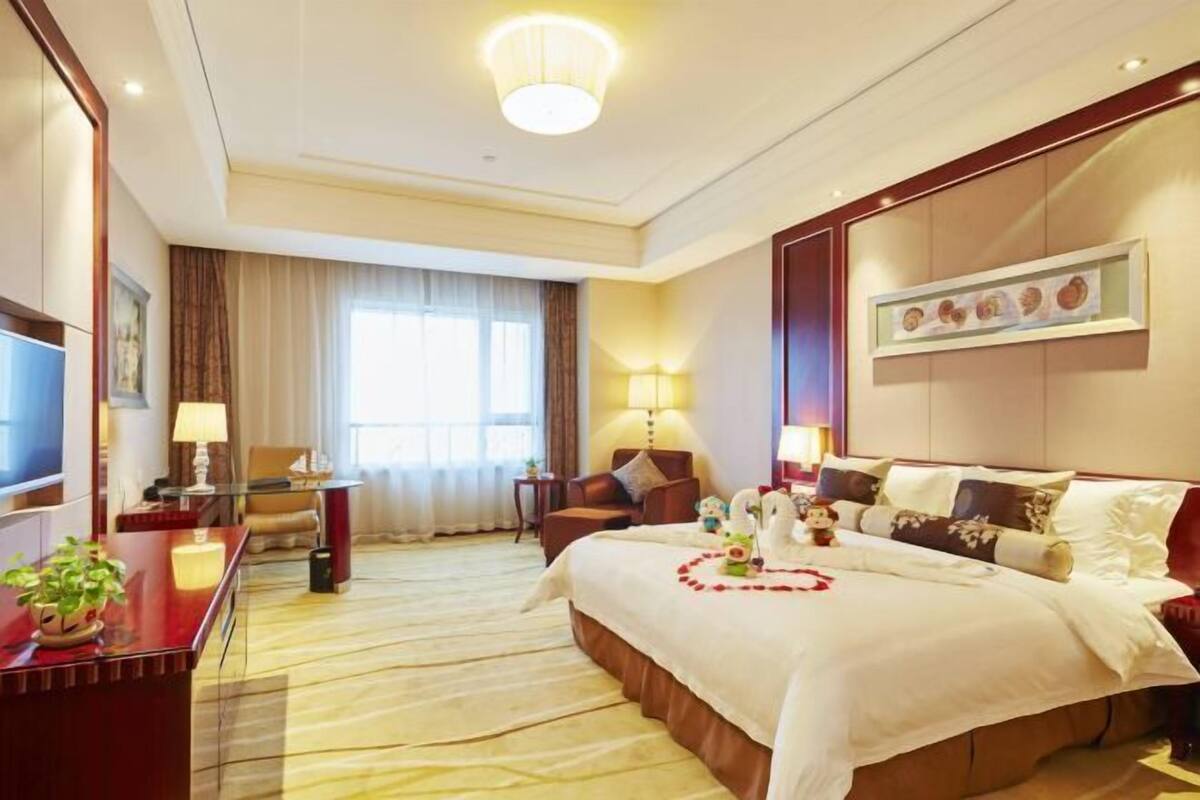 Ein Hotelzimmer mit einem großen Bett, einem Schreibtisch mit Lampe, einem Sessel, einem Fernseher und einem Fenster mit Vorhängen.
