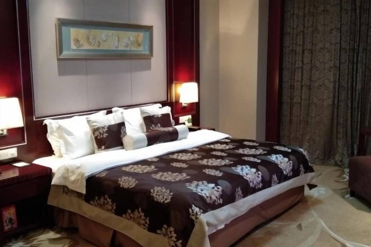 Ein Hotelzimmer mit einem großen Bett, zwei Nachttischen, einem Beistelltisch, einem gerahmten Bild an der Wand und einem Vorhang.