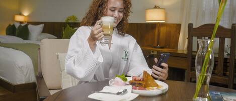 A person seated at a wooden table with a plate of food, a glass of beverage, and a smartphone.