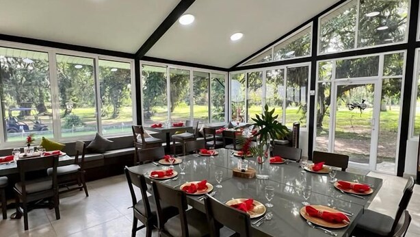 A spacious dining area with a large table set for six, surrounded by chairs. The room features large windows offering a view of the outdoors. - Roaring River Resort (Belmopan)