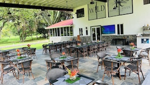An outdoor dining area with multiple tables and chairs, a stone wall, and a view of the surrounding greenery. - Roaring River Resort (Belmopan)