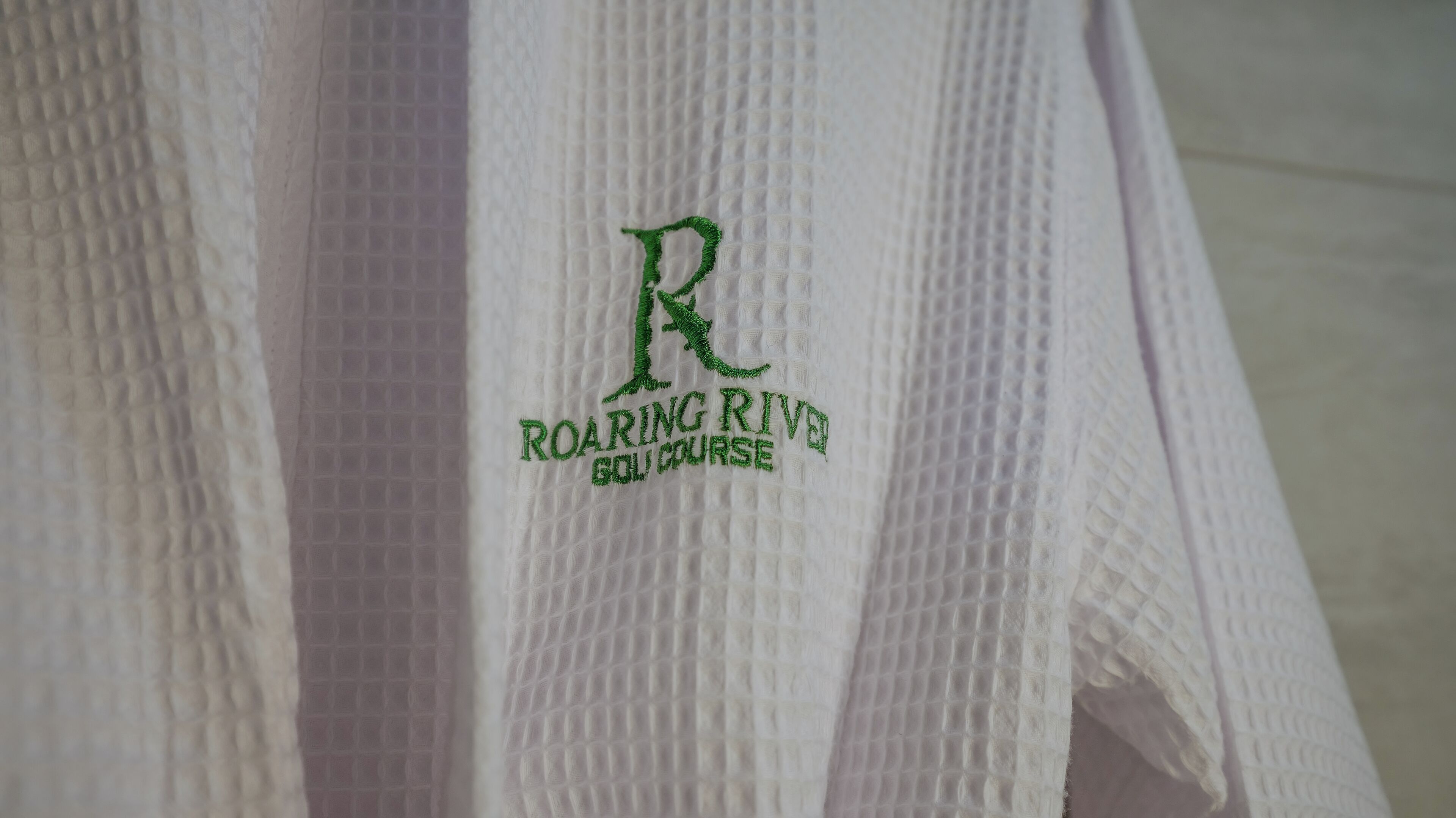 Ein weißes Handtuch mit aufgesticktem Text: „ROARING RIVER GOLF COURSE“.