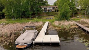 A boat docked at a wooden pier extending into a body of water, surrounded by lush greenery and a building in the background. - Gorgeous Waterfront Retreat (Pequot Lakes)