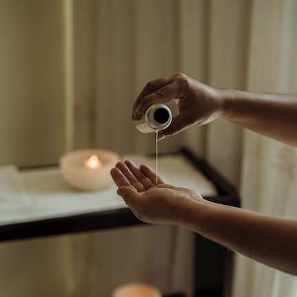 A hand holding a small object, possibly a candle, with a lit candle in the background.