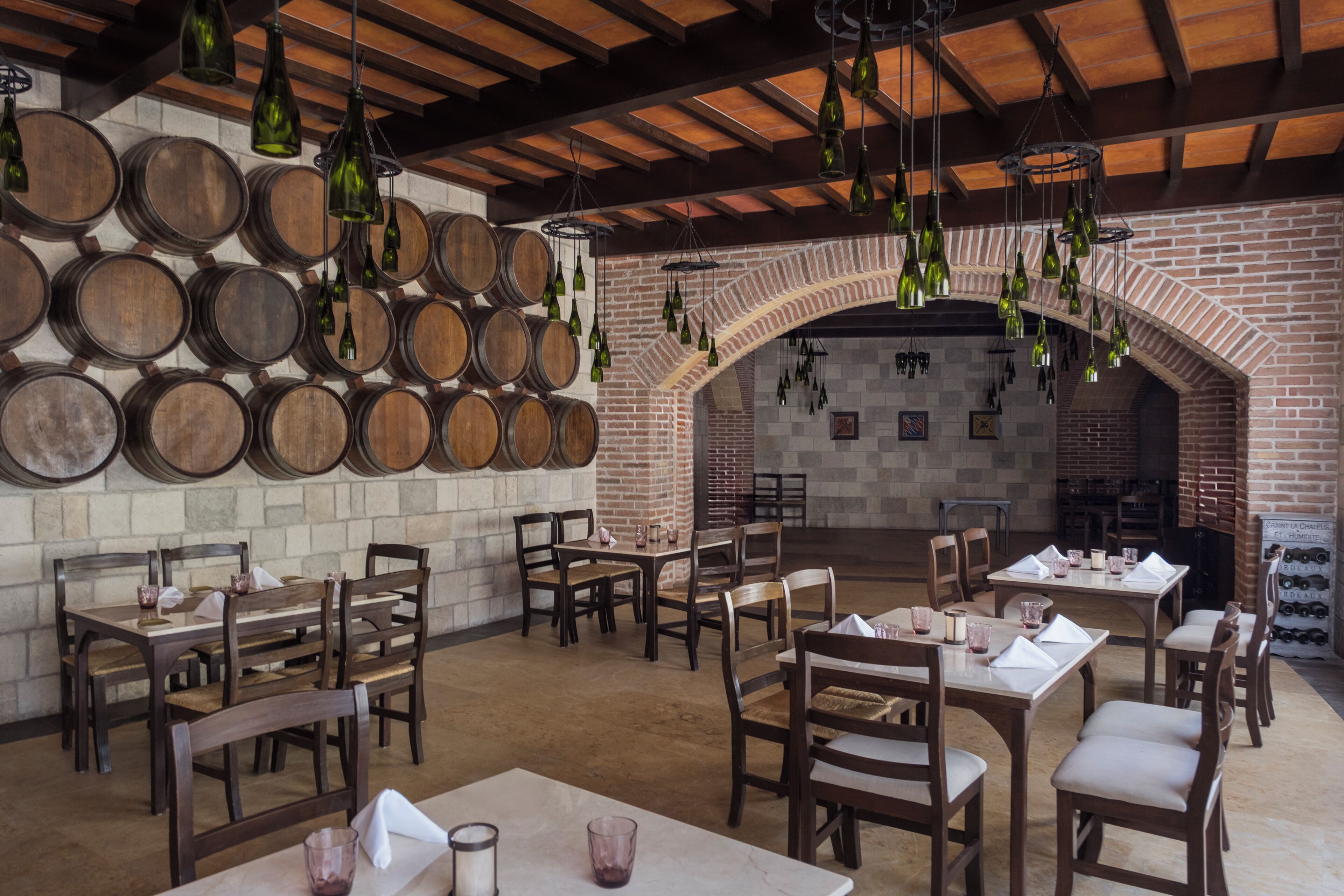 Un restaurante con mesas y sillas de madera, pared de ladrillo y botellas de vino colgando.