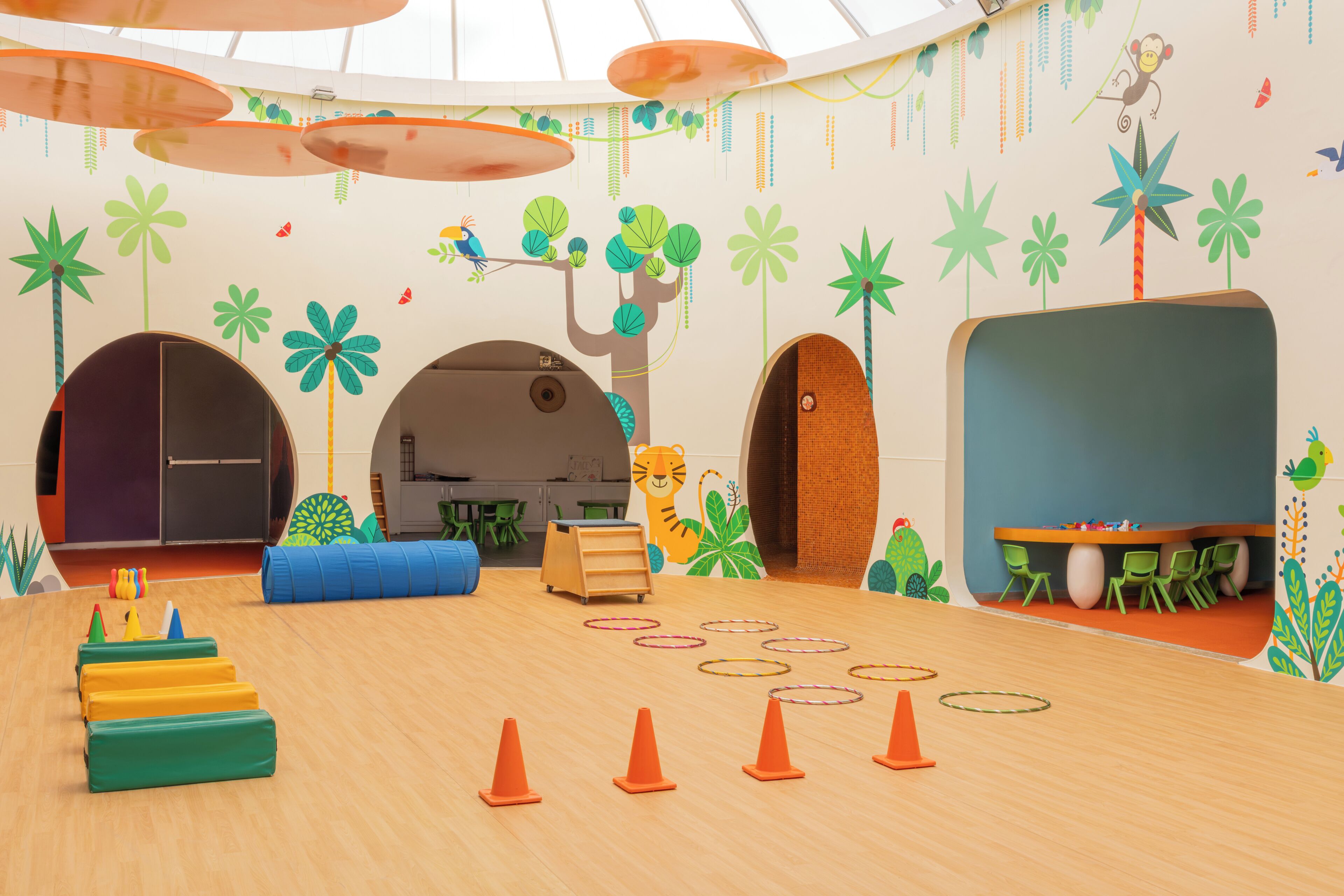 A children's playroom with a wooden floor, colorful play mats, and a large mural with trees and animals.