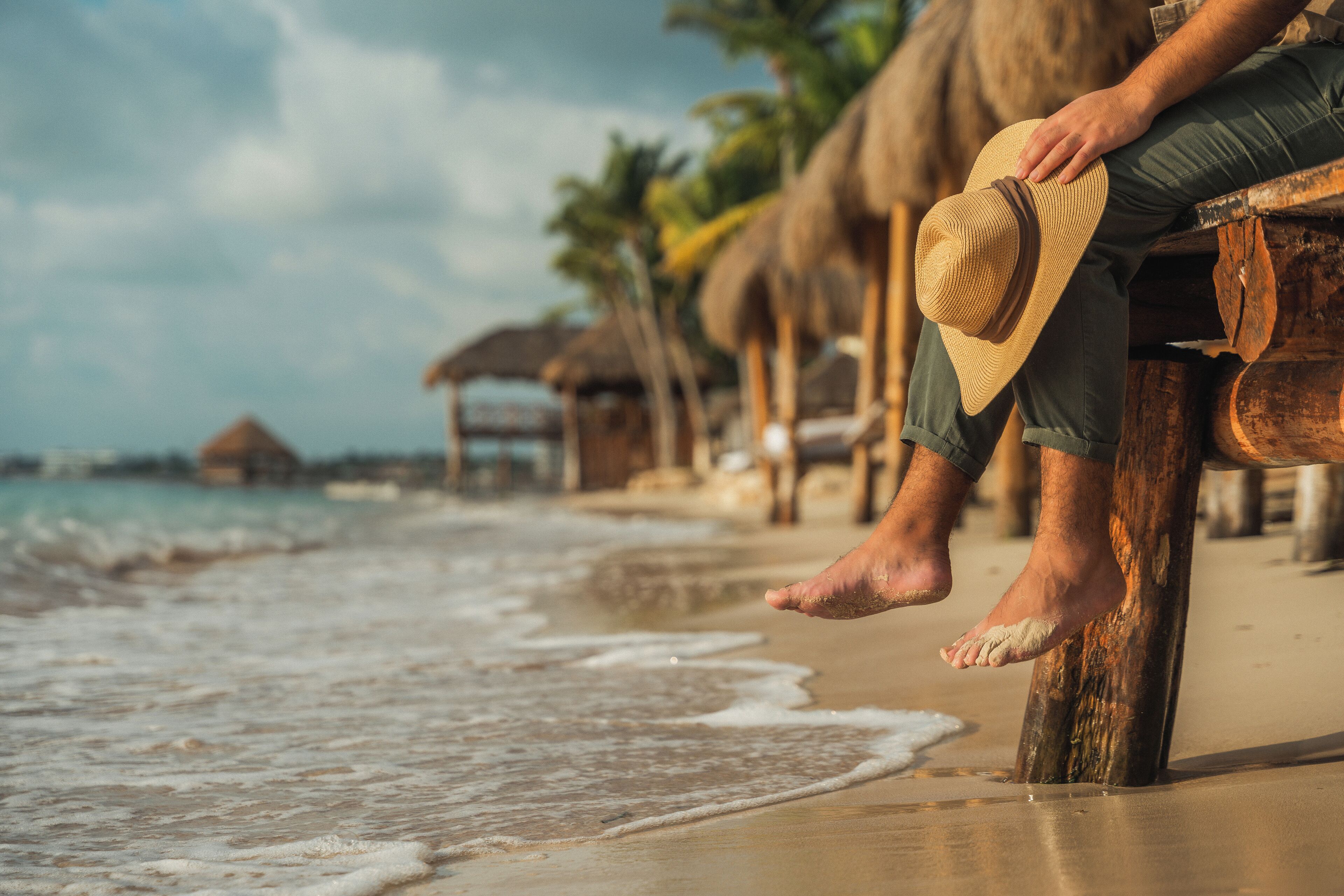Una persona sentada en un banco de madera junto a la playa, sosteniendo un sombrero de paja.