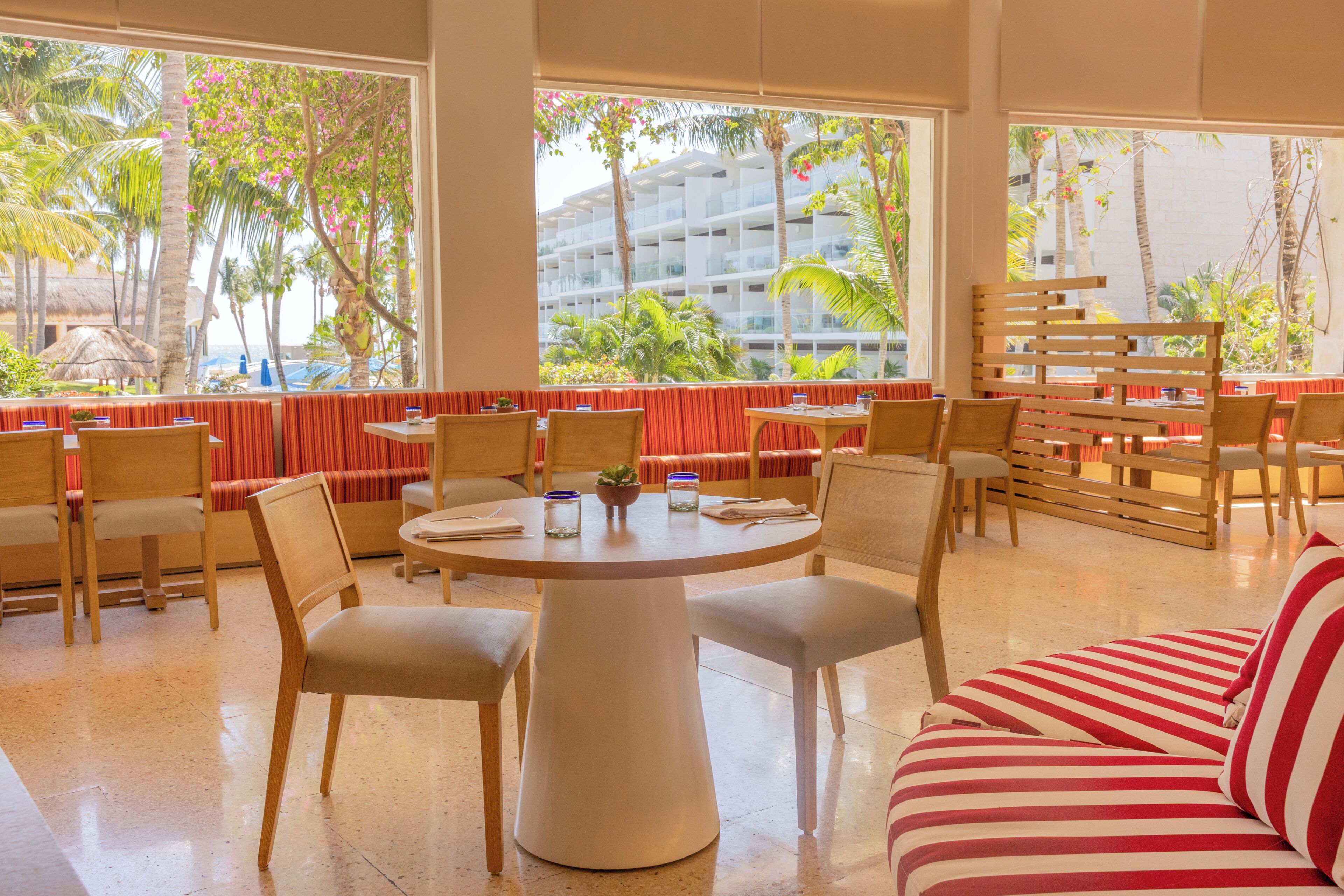 A hotel room with a round dining table, chairs, a striped sofa, and large windows with a view of palm trees and a building.
