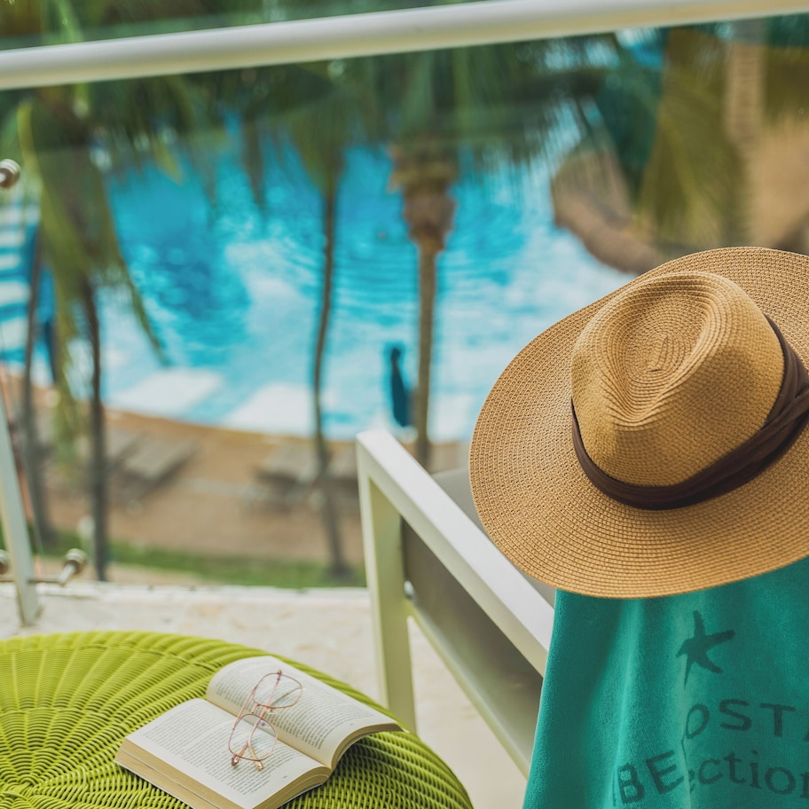 A balcony with a view of a pool, a green woven ottoman, a straw hat, and a towel with text.
