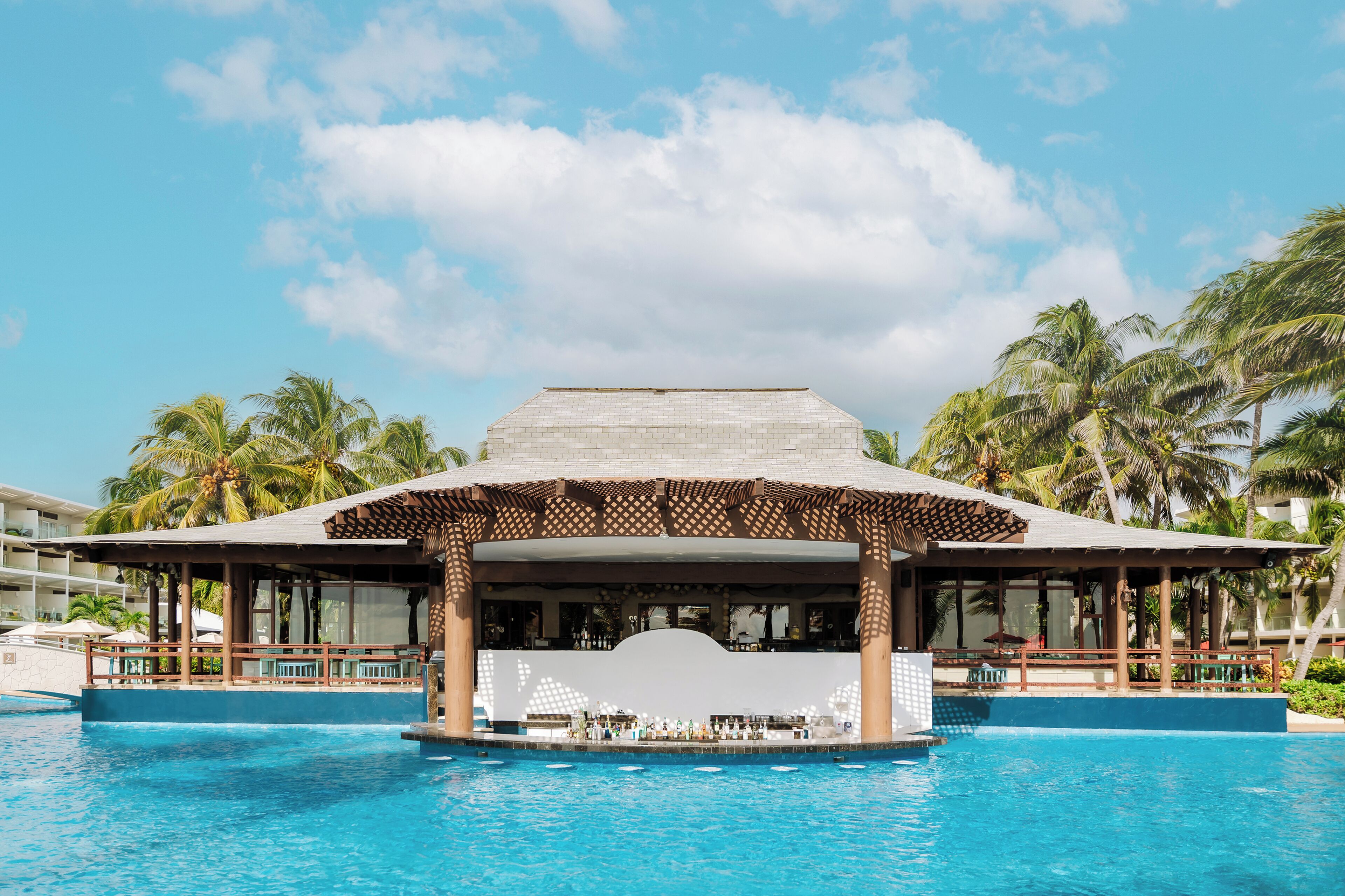 A resort pool area with a covered bar and lounge area, surrounded by palm trees and a clear sky.