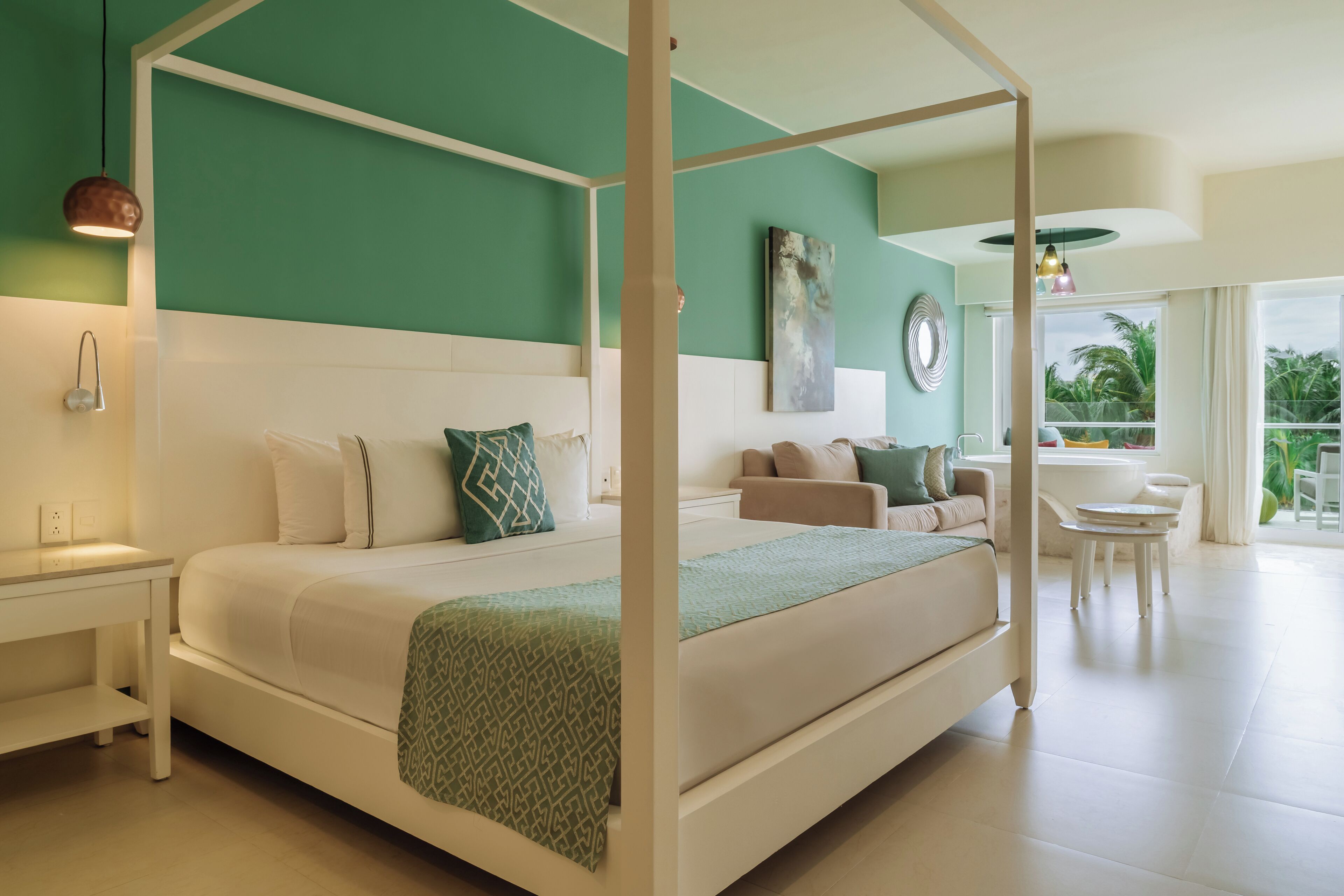 Un lit à baldaquin avec une tête de lit blanche, un plaid vert et un coussin à motifs. On y trouve une table de chevet blanche avec une lampe, un canapé beige avec des coussins et une petite table ronde avec des chaises. Un grand tableau abstrait orne le mur, et une fenêtre offre une vue sur des palmiers.