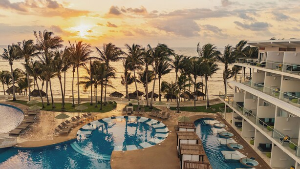 Een resort met een groot zwembad, ligbedden en uitzicht op de oceaan bij zonsondergang.