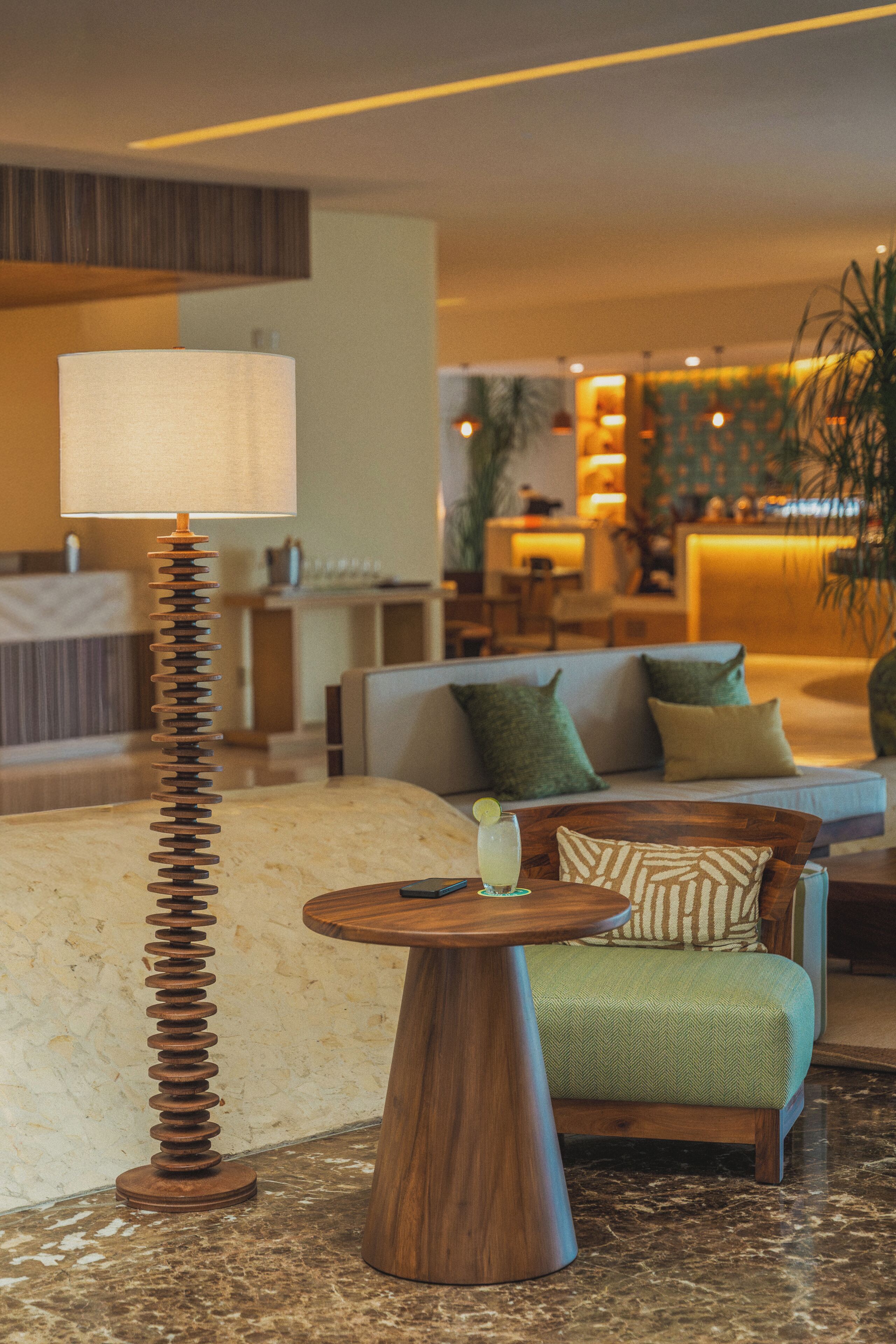Um lobby moderno com uma luminária única, um sofá com almofadas, uma mesa redonda pequena e uma área de bar ao fundo.