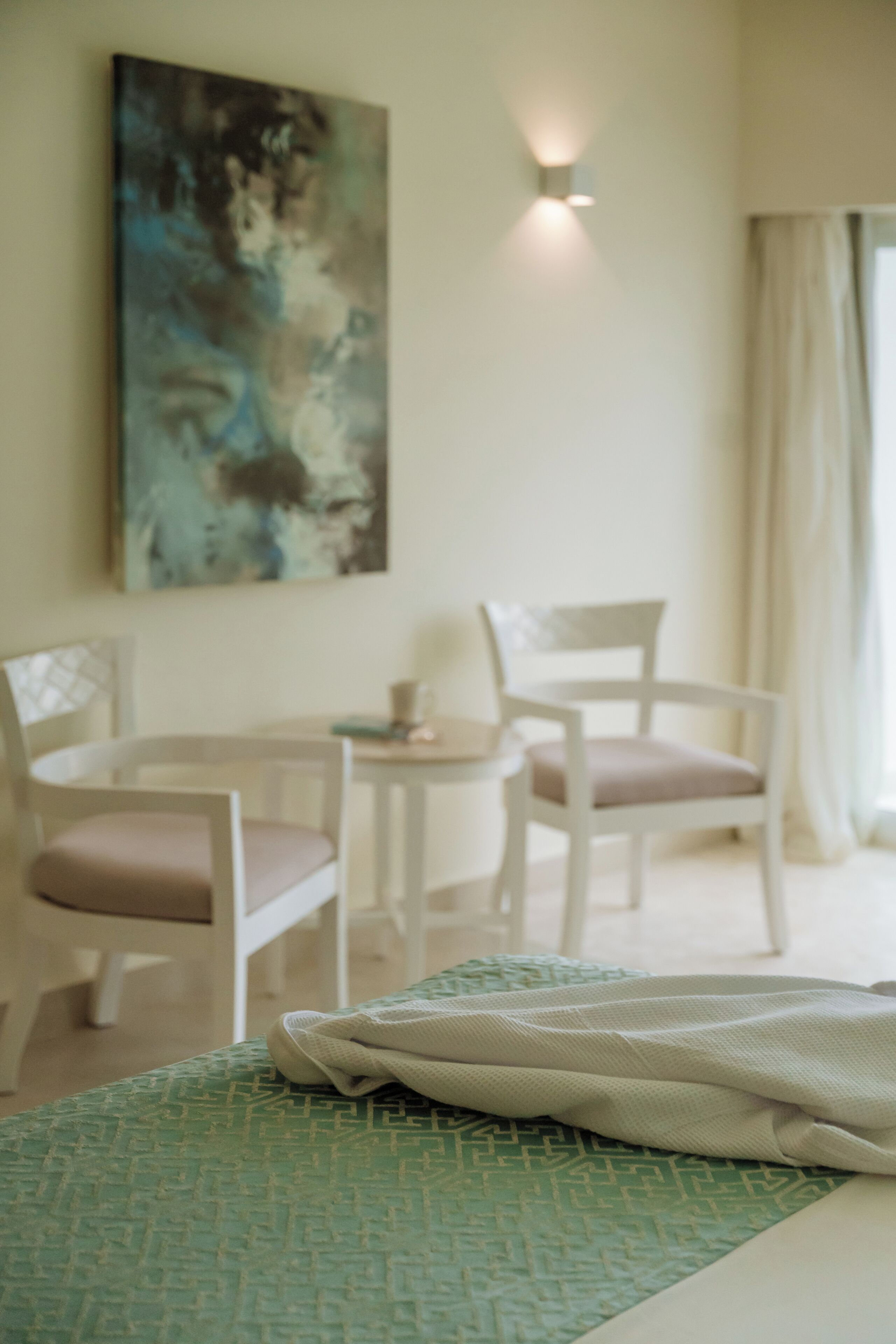Quarto de hotel com cama, duas cadeiras, uma mesa pequena e um quadro na parede.