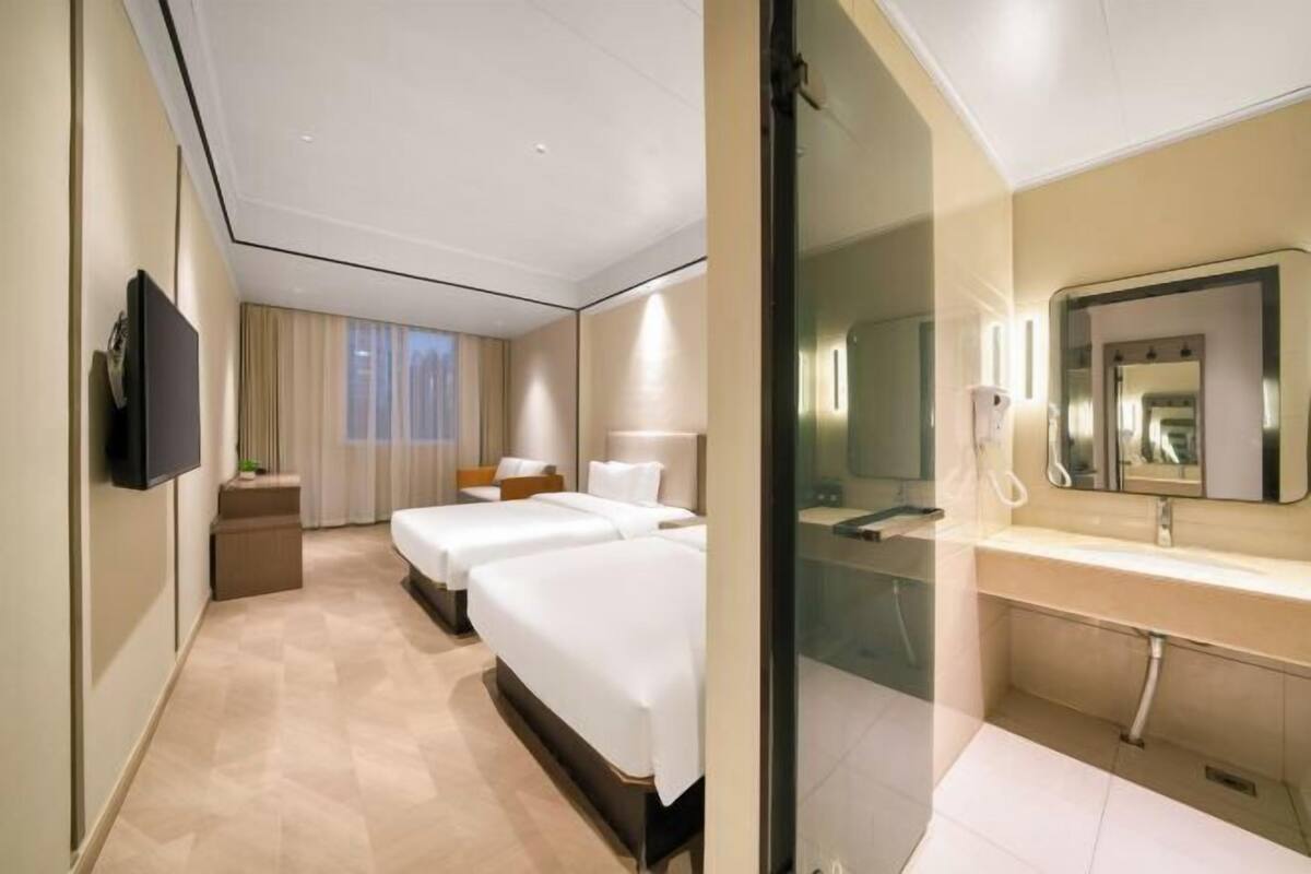 A hotel room with two beds, a TV, a bathroom with a mirror and sink, and a window with curtains.