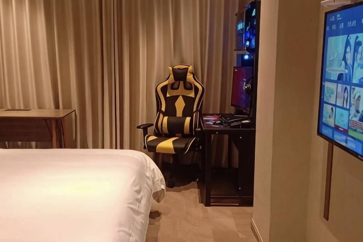 A hotel room with a bed, a gaming chair, a desk with a computer, and a television displaying a game.