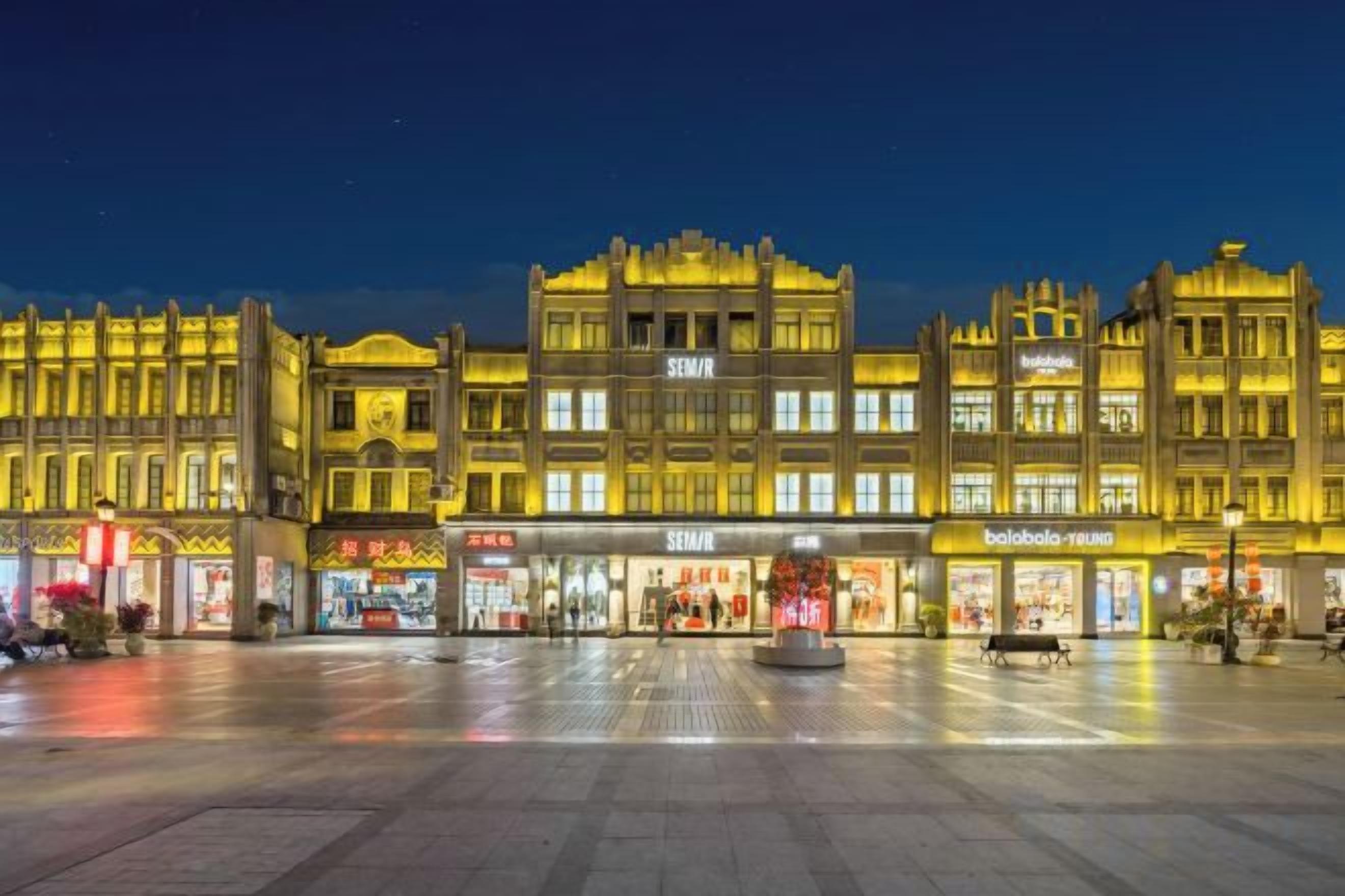 Un edificio grande e iluminado con varias tiendas, una plaza central abierta y una fuente.