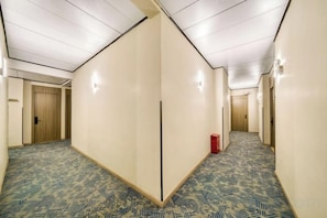 Een hotelgang met een kamerbreed tapijt, houten deuren en een rode prullenbak.