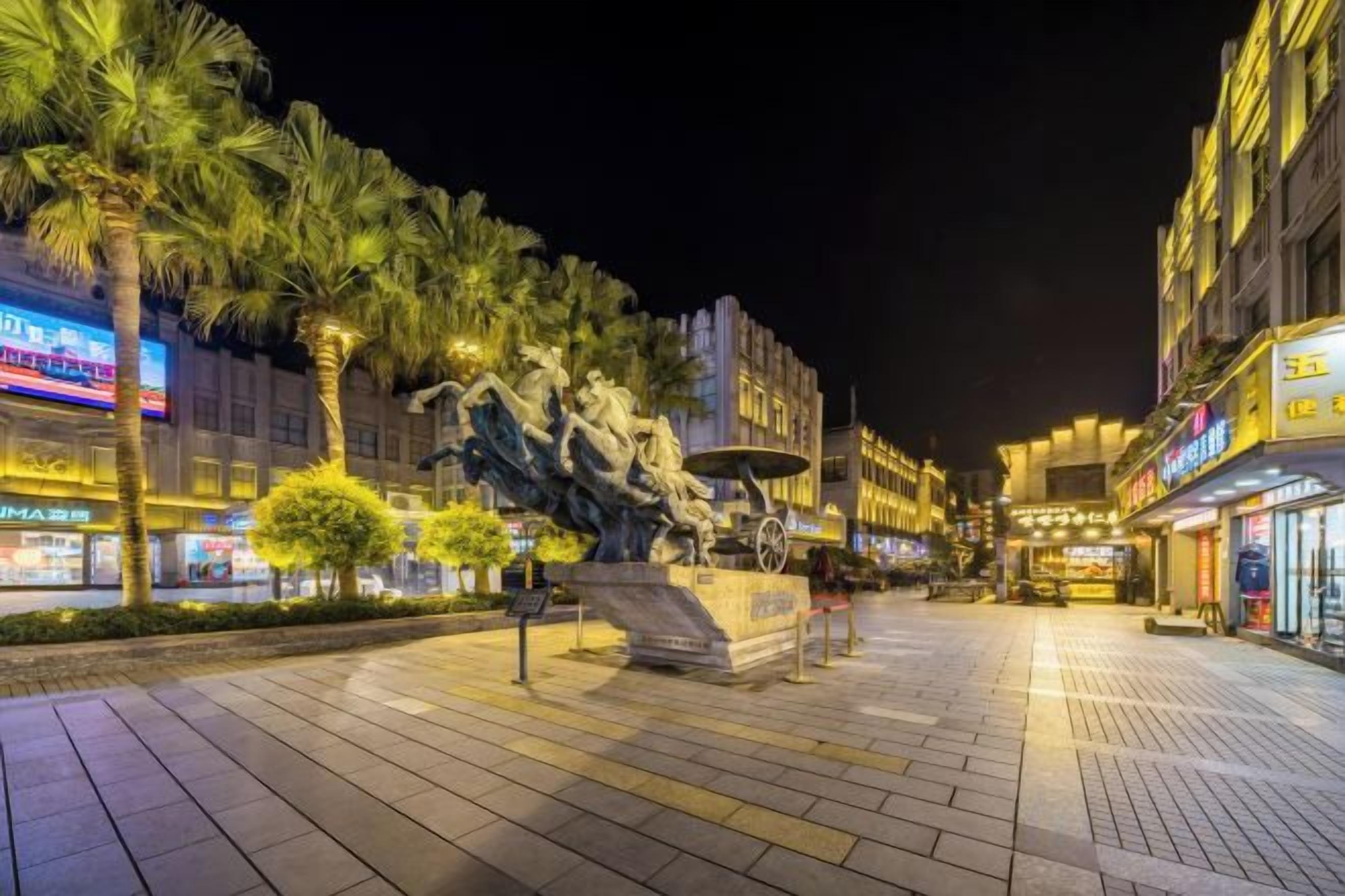 Una plaza urbana nocturna con una estatua central, palmeras y escaparates iluminados.