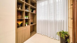 Una stanza con una libreria in legno piena di oggetti decorativi e piante, un'ampia finestra con tende leggere e una pianta in vaso sul pavimento.