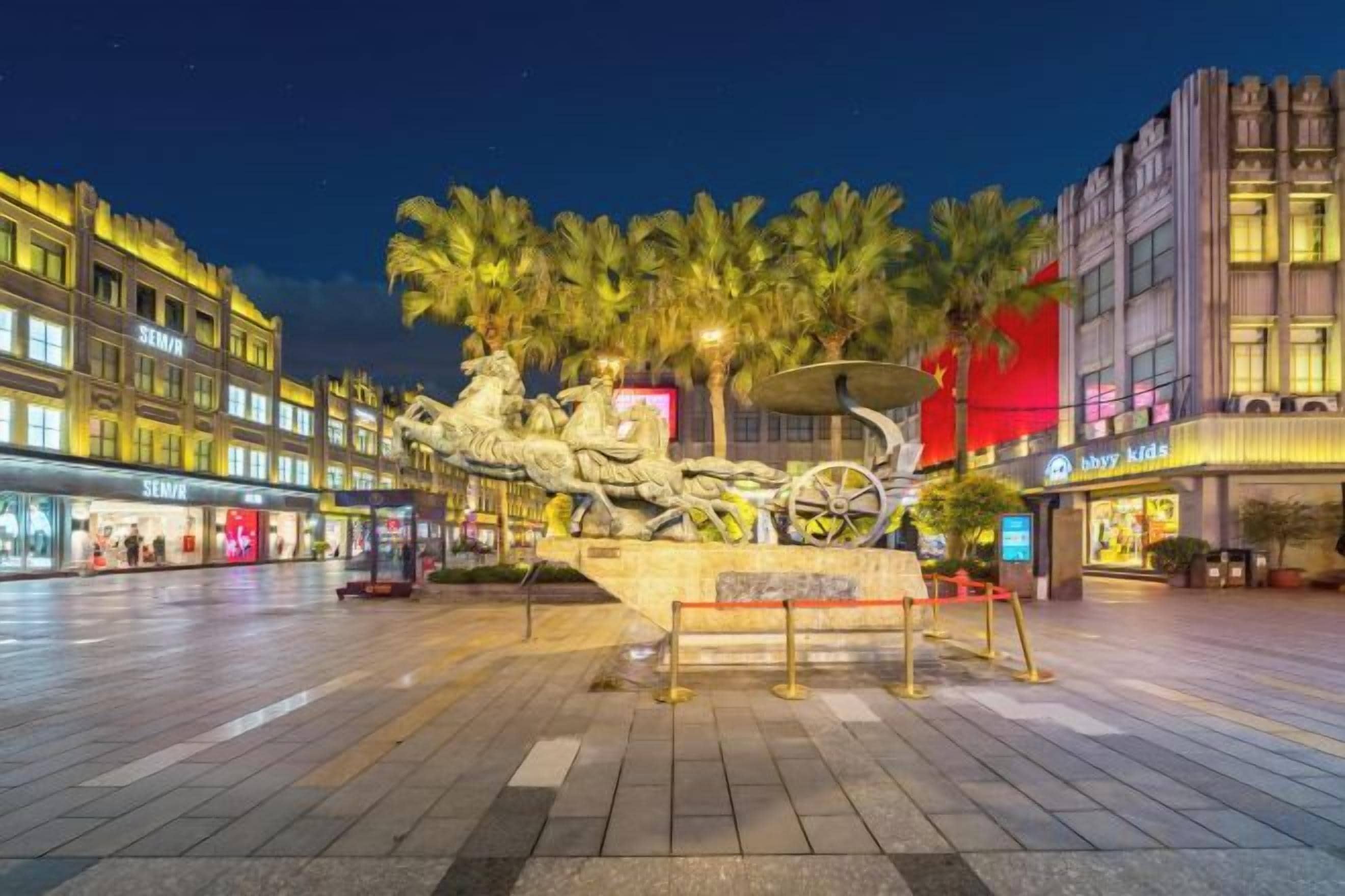A public square with a central sculpture, surrounded by palm trees and illuminated buildings.