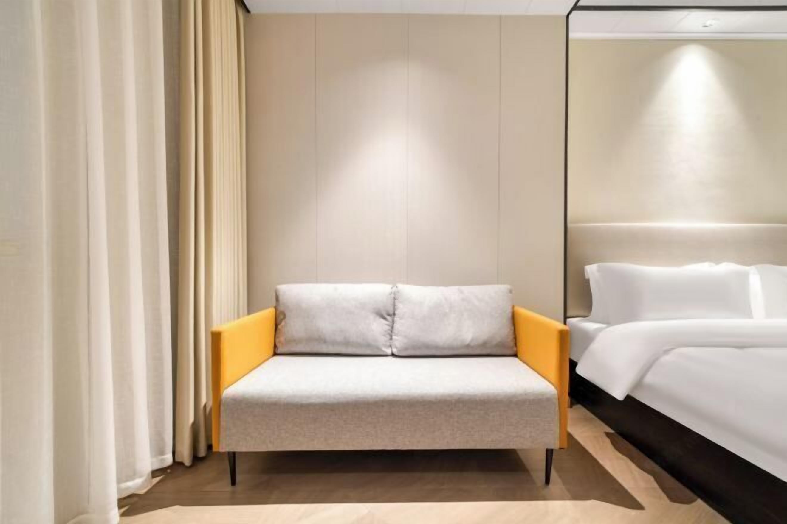 Une chambre d’hôtel moderne aux couleurs neutres, comprenant un canapé-lit recouvert d’un tissu gris clair, un plancher en bois et une grande fenêtre avec des rideaux.