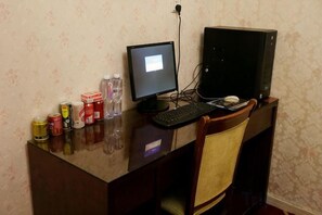 Un escritorio de computadora con un monitor, teclado y una computadora de torre. Hay varios objetos sobre el escritorio, incluyendo una botella, latas y una caja.