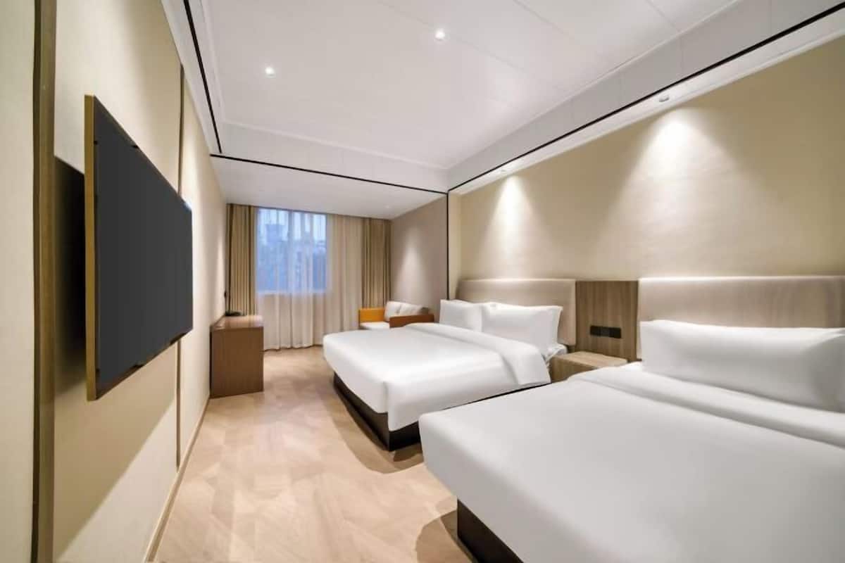 A hotel room with two beds, a flat-screen TV mounted on the wall, a bedside table with a lamp, and a window with curtains.