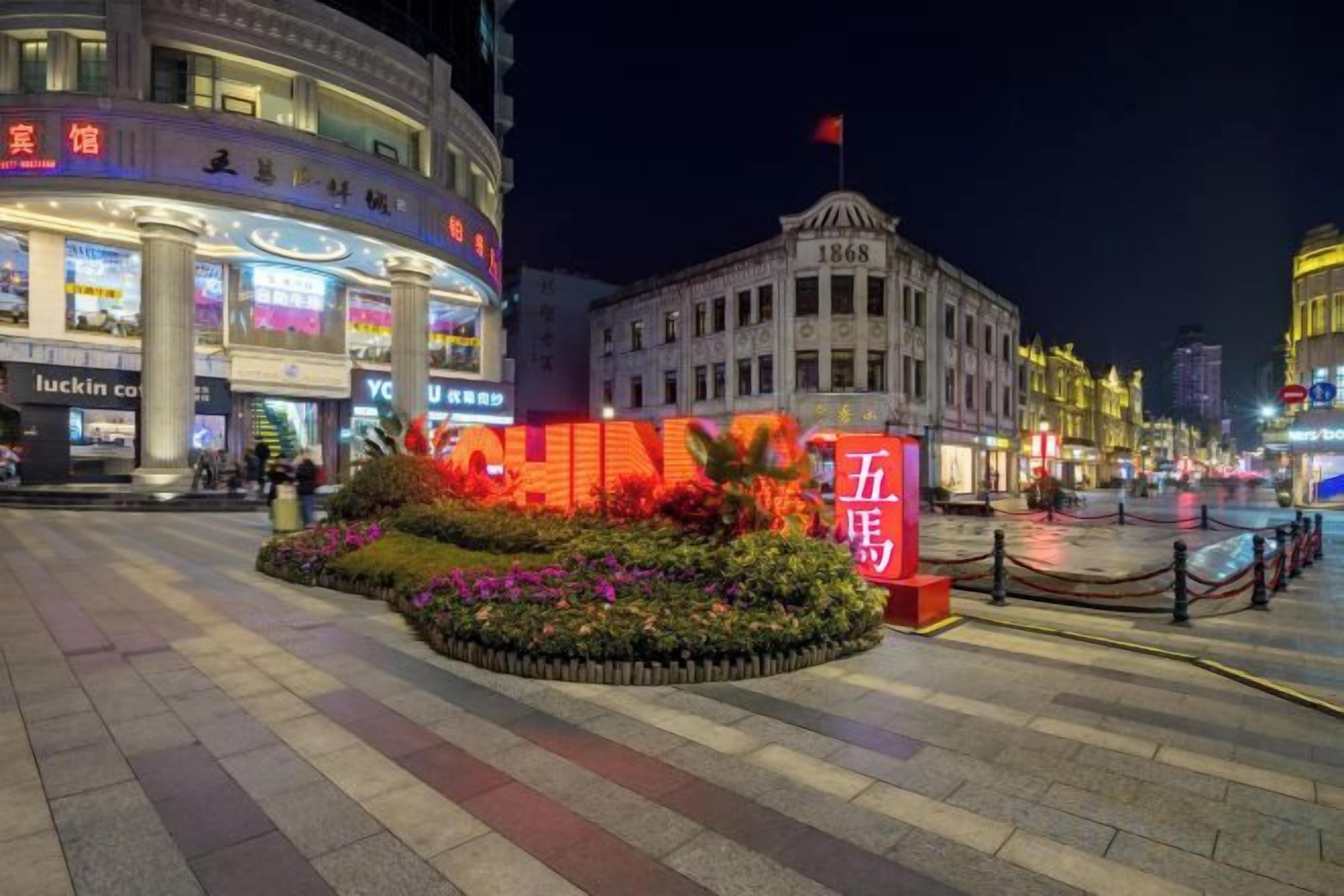 A well-lit urban square with a prominent red sign, a flower bed, and a historic building with a flag.