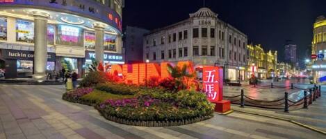 A well-lit urban square with a prominent red sign, a flower bed, and a historic building with a flag.