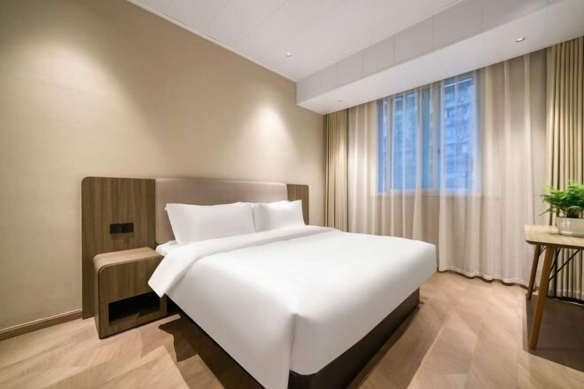 A modern hotel room with a large bed, a bedside table, a small desk with a plant, and a window with curtains.